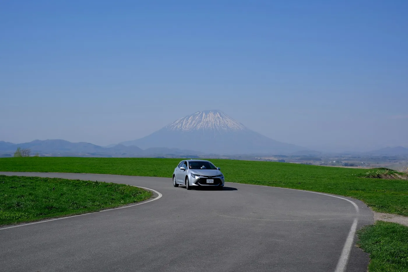 A car on a quiet rural road in Hokkaido with the snow covered cone of Mount Yotei behind it