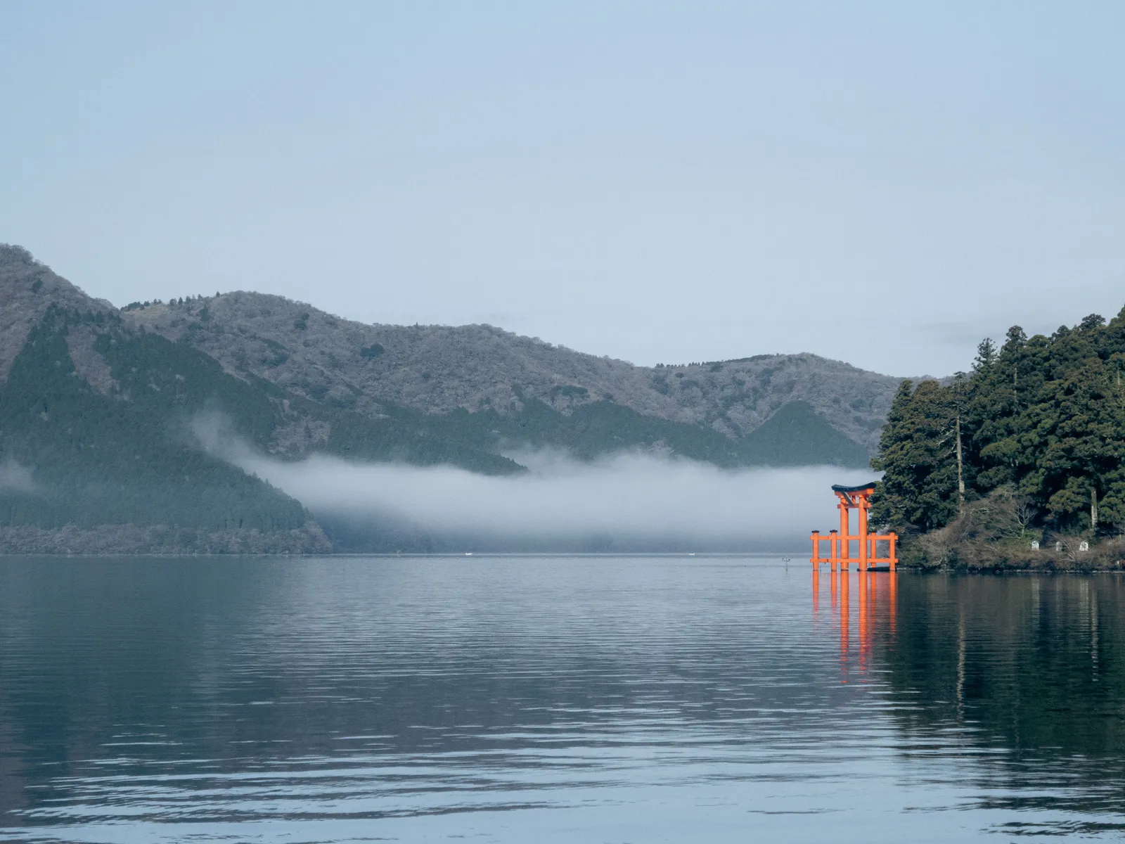 The red torii gate of Hakone shrine standing in the water of Lake Ashi with mountains behind