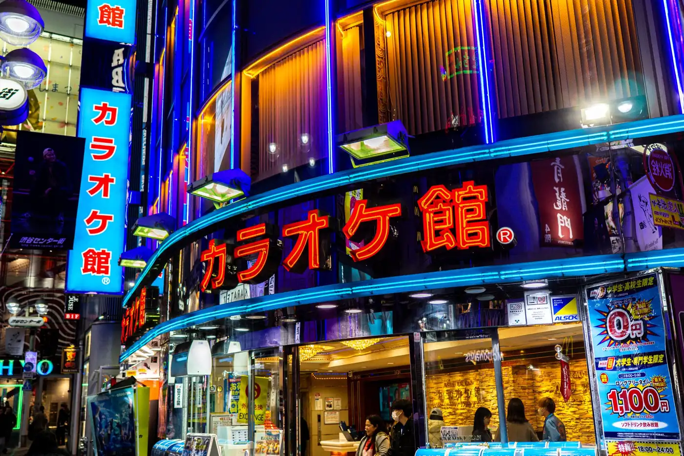 The neon lit street frontage of a karaoke building at night in Japan, with large illuminated signs in Japanese script above the entrance