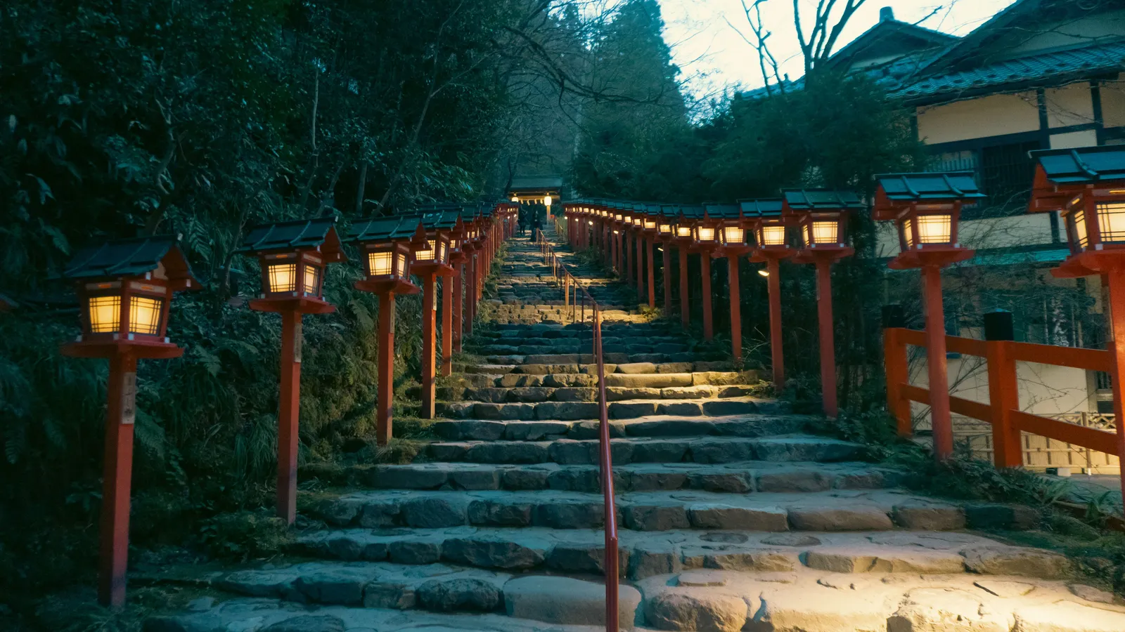 A stone stairway lined with red lanterns and torii posts climbing into trees at a shrine in Kyoto, Japan