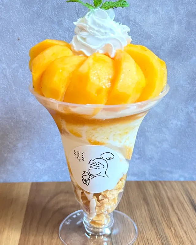 A tall glass with mango slices, whipped cream and a mint leaf, against a light background