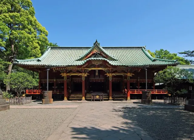 Vermilion shrine hall with a curved roof above a stone courtyard, green trees behind