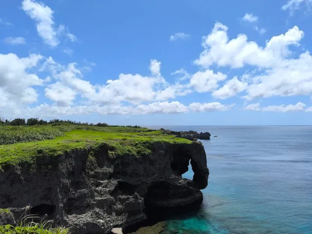 A grass topped rock cliff with an arch carved above the sea, and turquoise water below