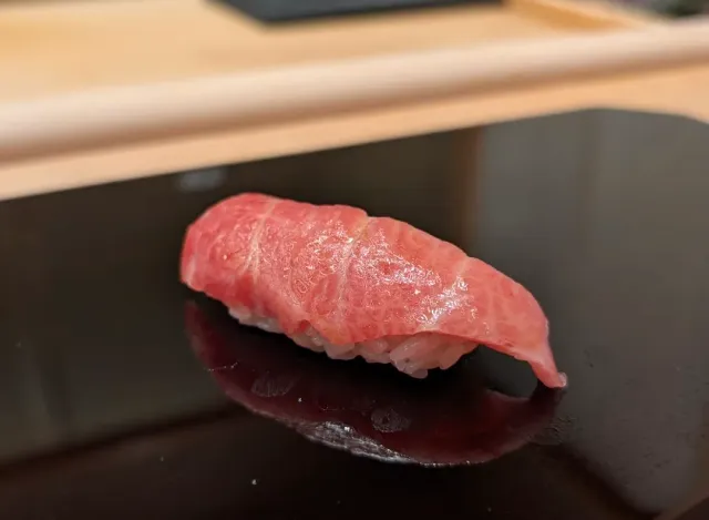 A single piece of rice wrapped in a pink slice of fish, on a dark glossy lacquer surface