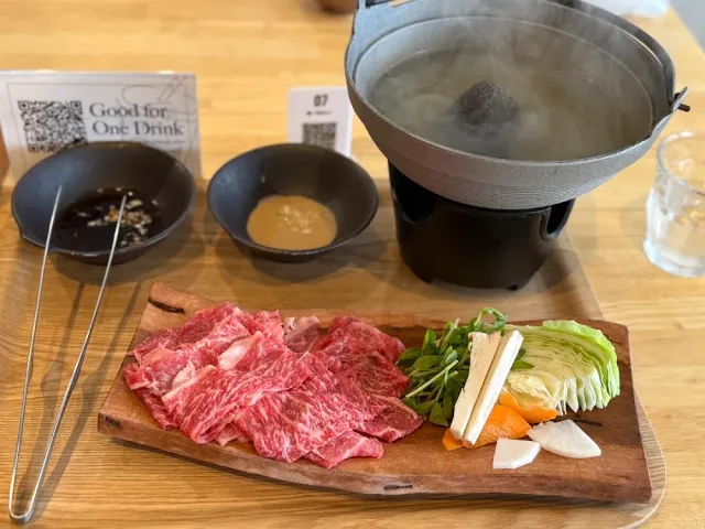 A steaming pot on a burner, a wooden board with marbled slices of meat, vegetables and dipping bowls