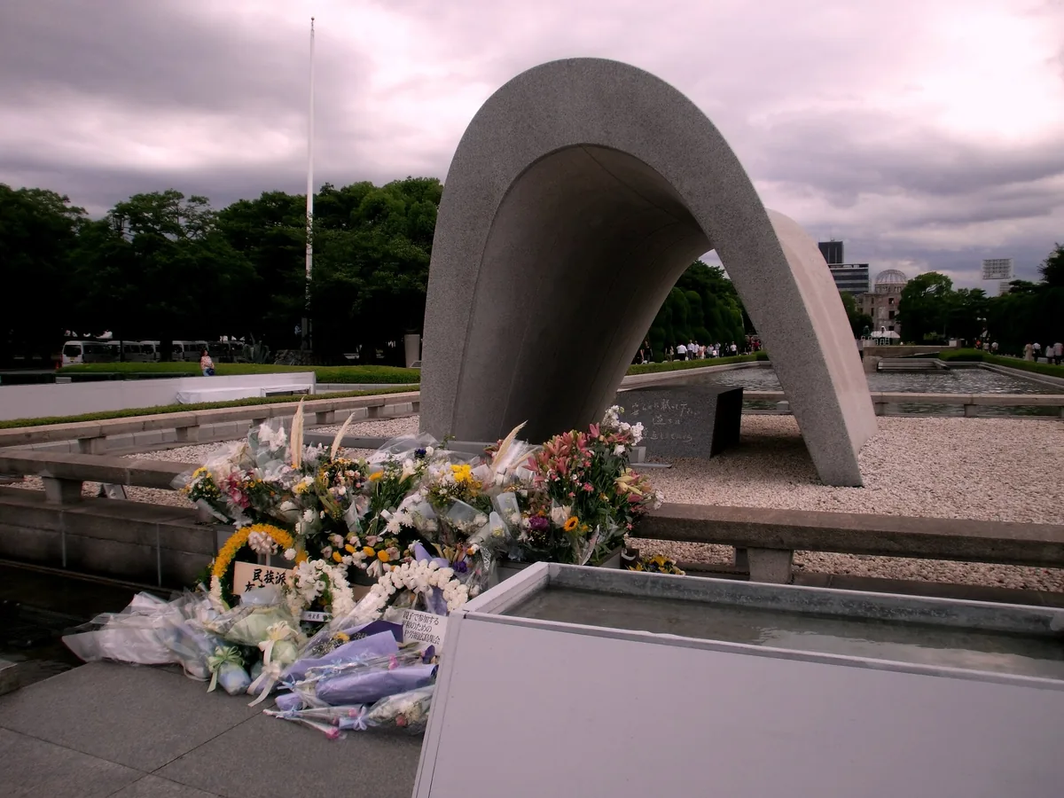 The concrete arch of the Memorial Cenotaph in Hiroshima Peace Memorial Park, flower wreaths at its base and the Atomic Bomb Dome beyond the trees behind it