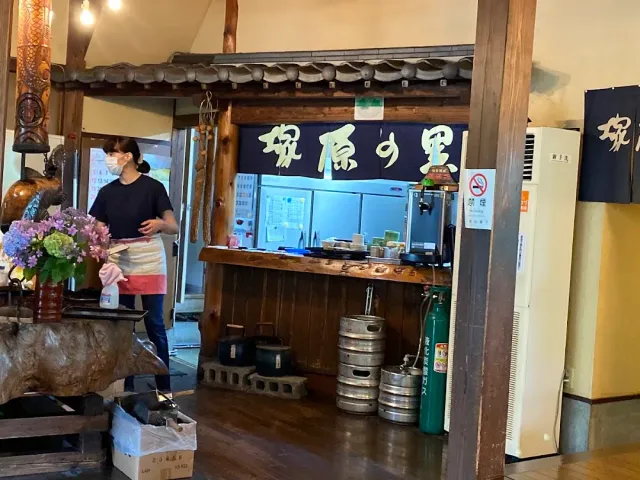 A worker standing by a wooden counter under a tiled eave, with a blue cloth curtain and Japanese lettering