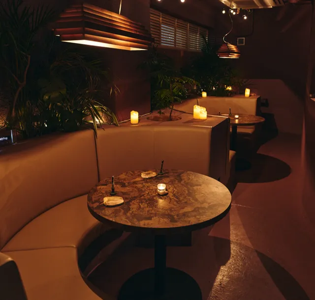 Curved sofas around marble tables, lit candles and plants in dim lighting