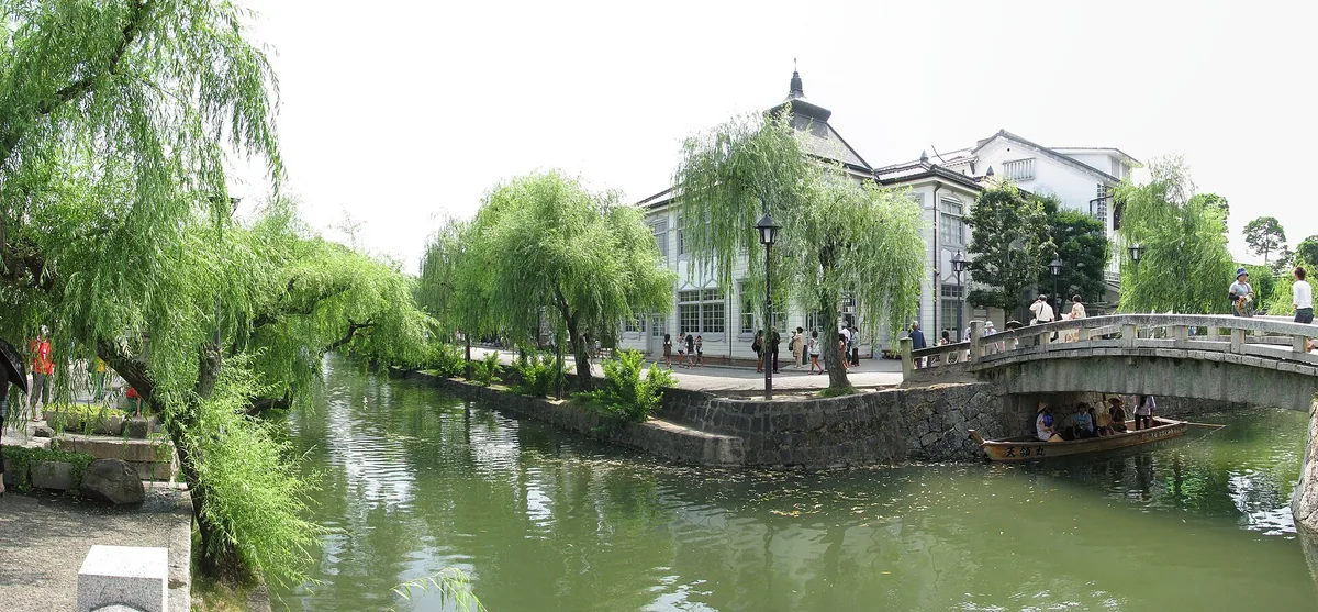 The Bikan canal in Kurashiki with weeping willows, historic white storehouse buildings, and a small tour boat with passengers on the water