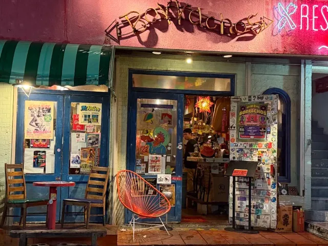 A frontage at night with lit lettering, a striped awning, blue doors with stickers and an orange chair