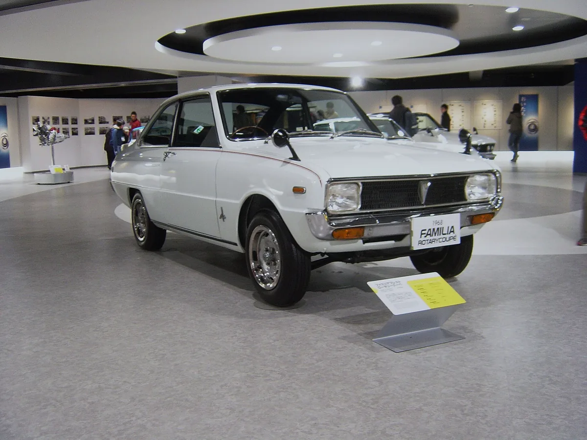 A white vintage Mazda coupe on a plinth in a lit showroom hall