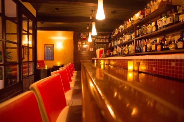 A long wooden counter with red stools and a wall of bottles in warm light