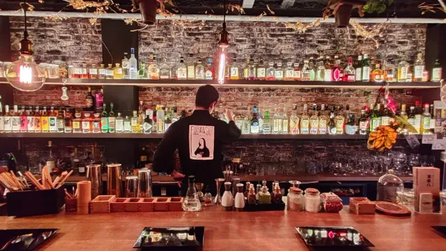 A bartender standing at a brick wall with shelves holding dozens of bottles