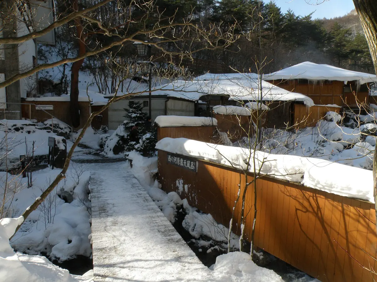 The brown wooden fence of the Sainokawara Rotenburo in Kusatsu under snow, with a bridged path to the entrance