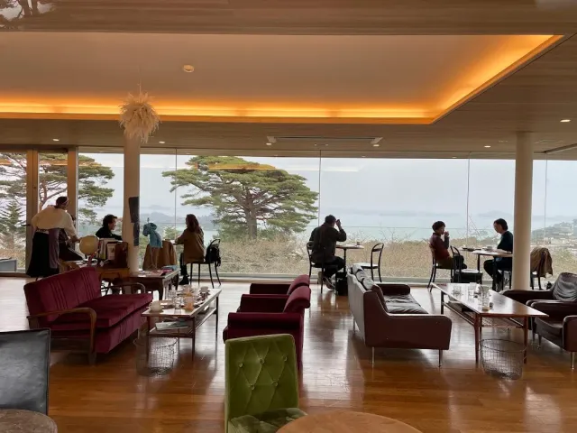 A seating lounge with sofas and large windows, and beyond them pine trees and a grey bay