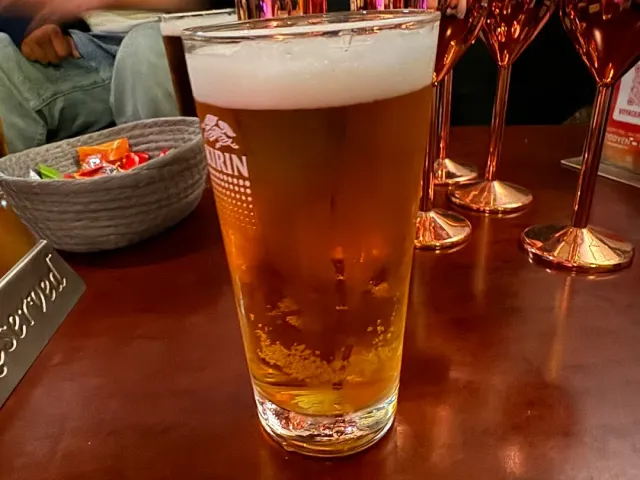 A glass of beer with foam on a wooden counter beside metal goblets and a bowl of sweets