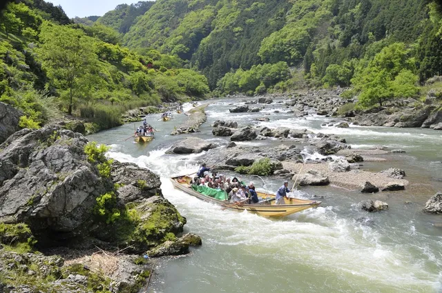 A flat wooden boat with passengers floats between rocks on a river, in a forested gorge