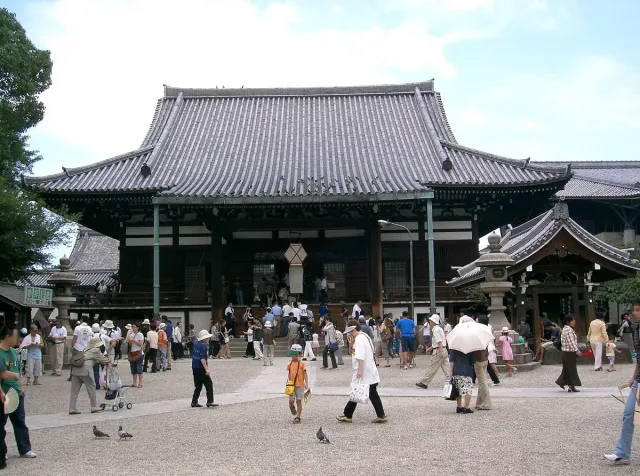 A wooden temple hall with a dark tiled roof and visitors in the courtyard