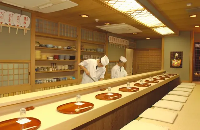 A long pale wooden counter with brown lacquer trays, and two cooks in white behind it