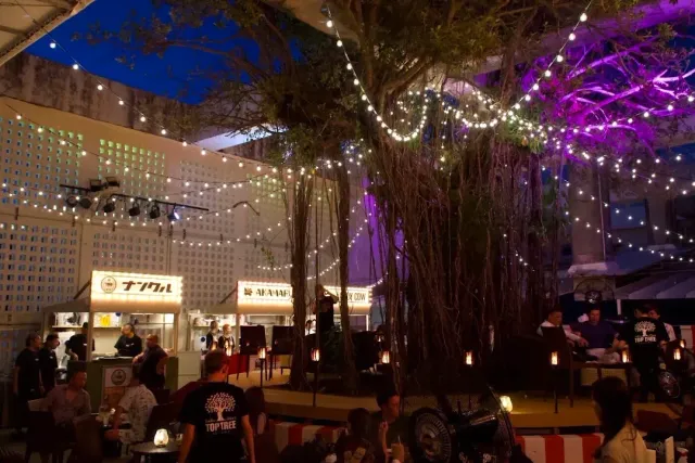 A night courtyard with a large tree, light strings, food stalls and people sitting