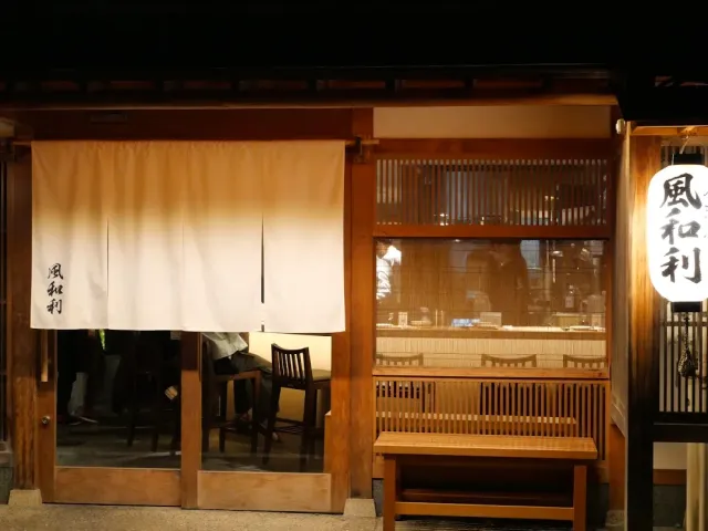 A frontage at night with a cream fabric curtain, glass doors, a wooden bench and a round lit lantern