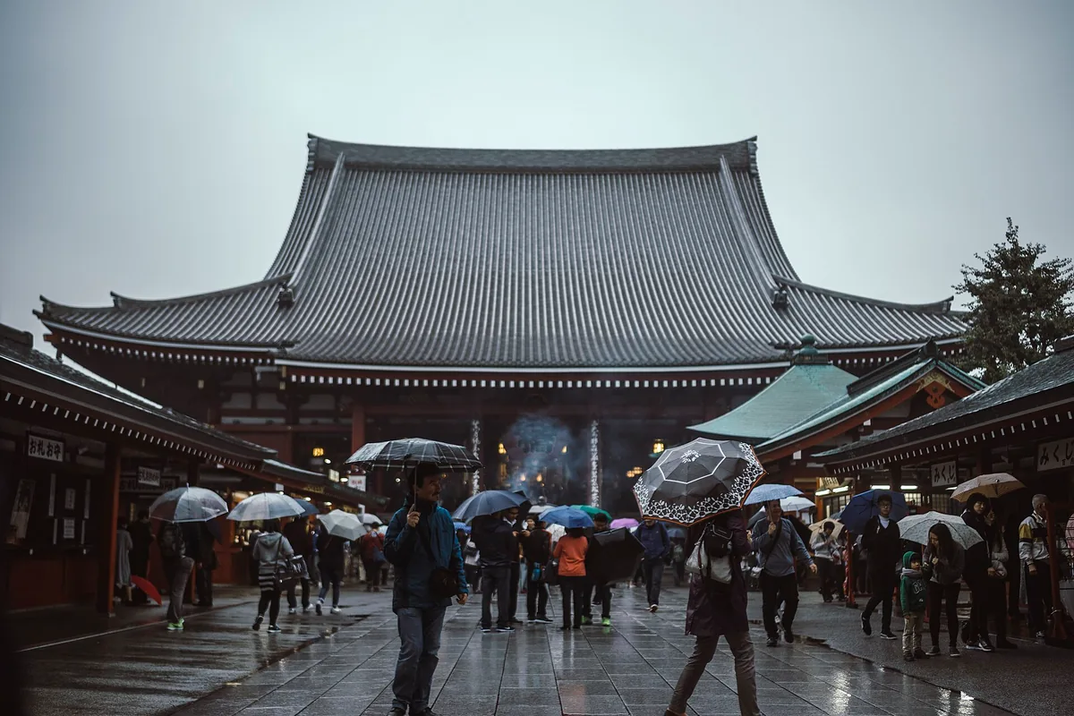A wooden temple hall with a curved tiled roof and visitors with umbrellas on a wet stone plaza