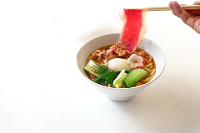 A bowl of ramen with green vegetables, as chopsticks lift a slice of meat