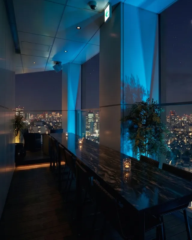A long table in blue lighting beside tall windows overlooking the city at night