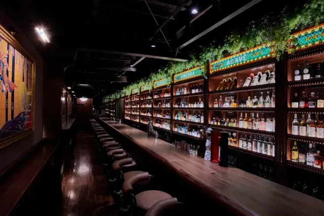 A long counter with a row of stools and a shelf wall full of bottles and greenery