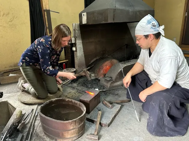 A smith in work clothes and a woman kneeling beside a forge with glowing metal