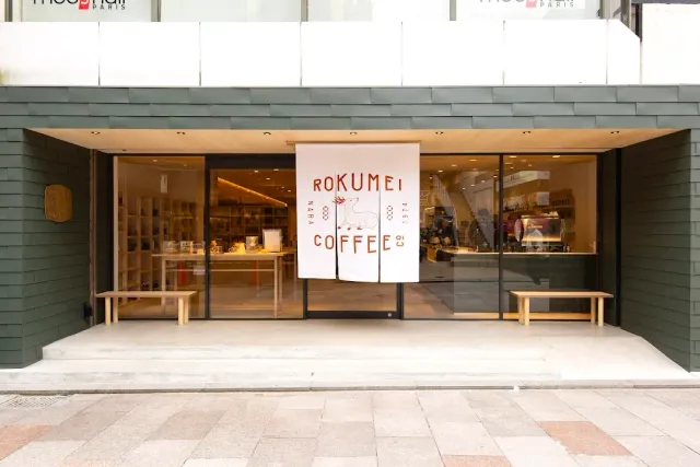 A shopfront with green walls, a hanging white cloth sign and wooden benches on both sides