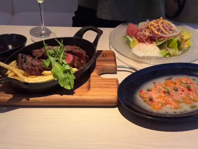 An iron pan with pieces of meat and fries, beside a salad plate and a fish dish