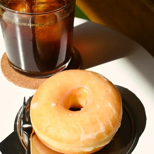 A glass of iced coffee on a cork coaster beside a glazed doughnut on a plate