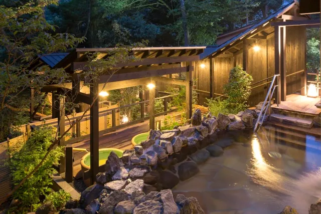 Bathing pools among rocks and a lit wooden building in a grove at dusk