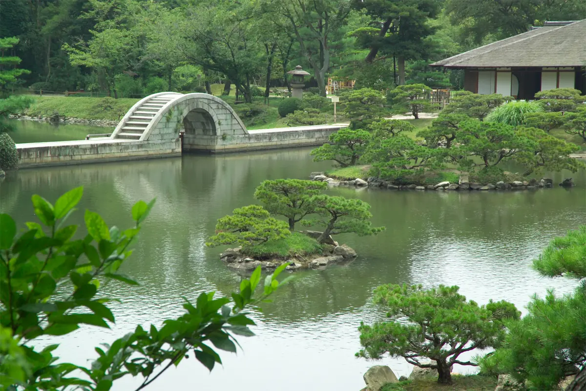 An arched stone bridge over a pond, framed by pines and greenery