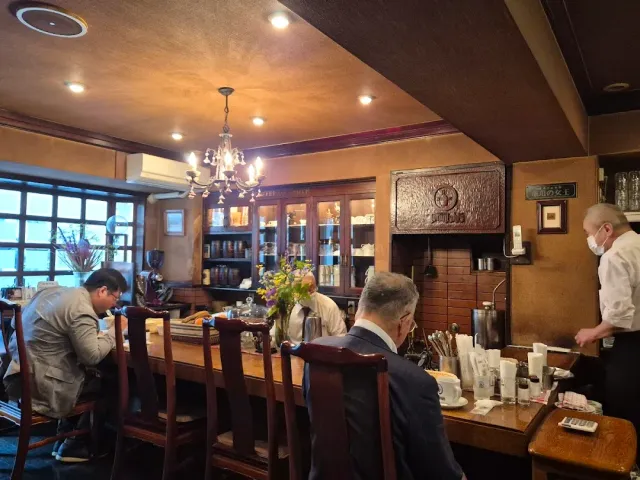 An old style room with a wooden counter, a chandelier, shelves of wares and seated customers