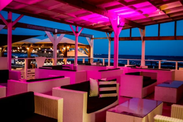 Wicker sofas under a wooden pergola in pink light, with the sea beyond at dusk