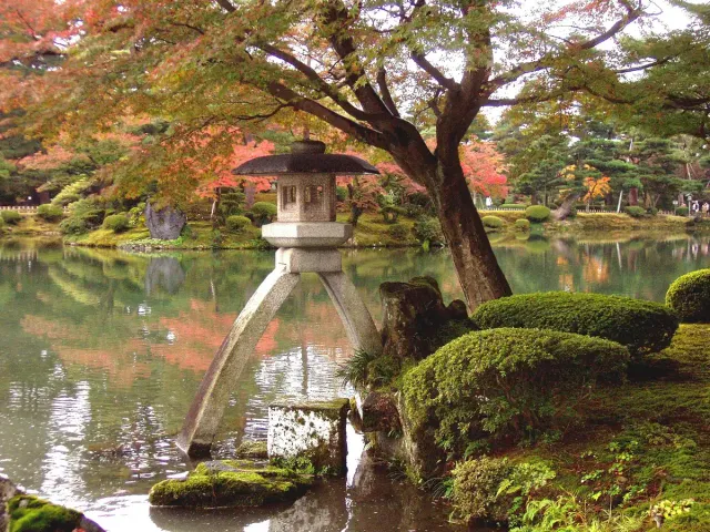 A stone lantern on slender legs at a pond edge, among trees and moss
