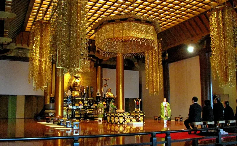 A gilded prayer hall with hanging golden canopies and an altar crowded with ritual objects, a monk in green robes on the polished wooden floor