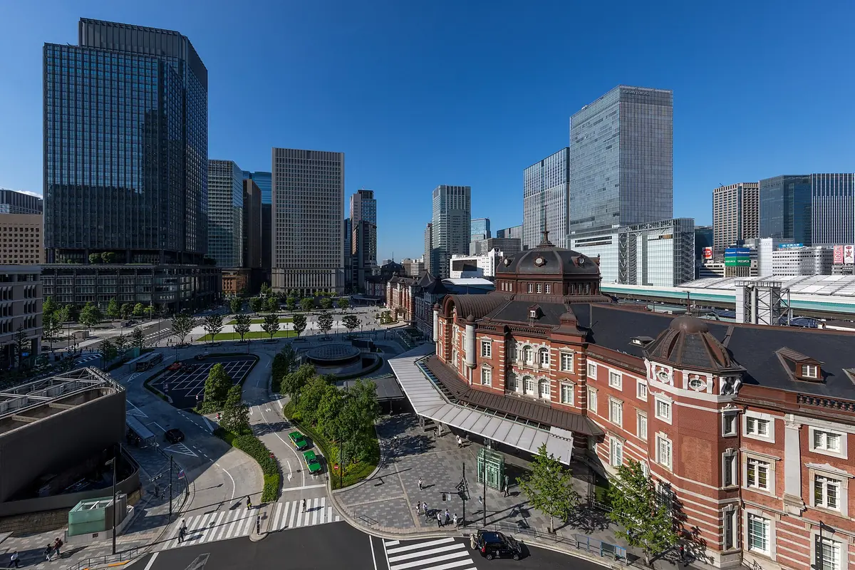 A red brick building with domes and a paved plaza in front, with glass towers under a blue sky