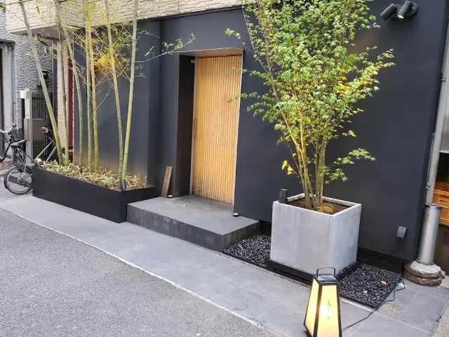 A dark frontage with a lit wooden door, bamboo and maple planters and a lantern on the pavement