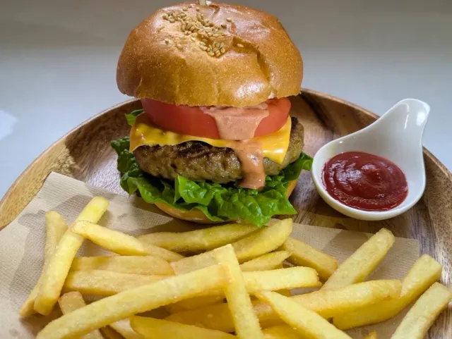 A hamburger in a sesame bun on a wooden plate, beside fried potatoes and red sauce