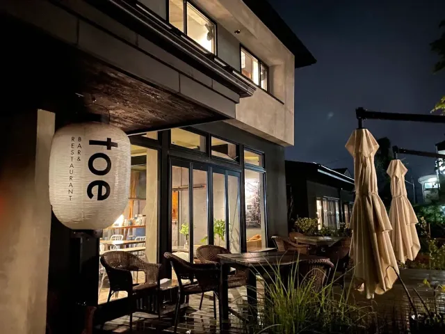 A facade lit at night with a large paper lantern, a terrace of chairs and closed parasols
