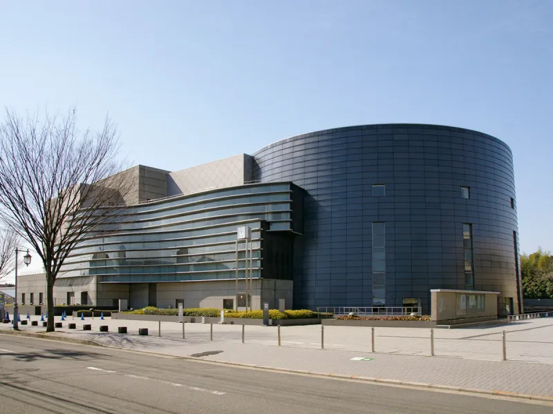 Rounded modern building clad in dark glass on a wide street, with bare trees in front