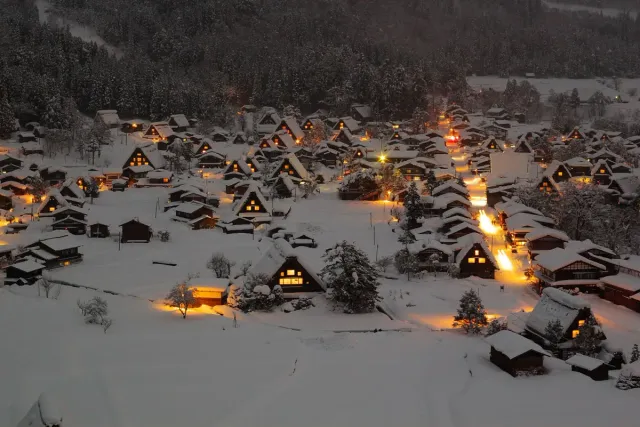 A village of steep thatched roofs lit up in the snow at dusk