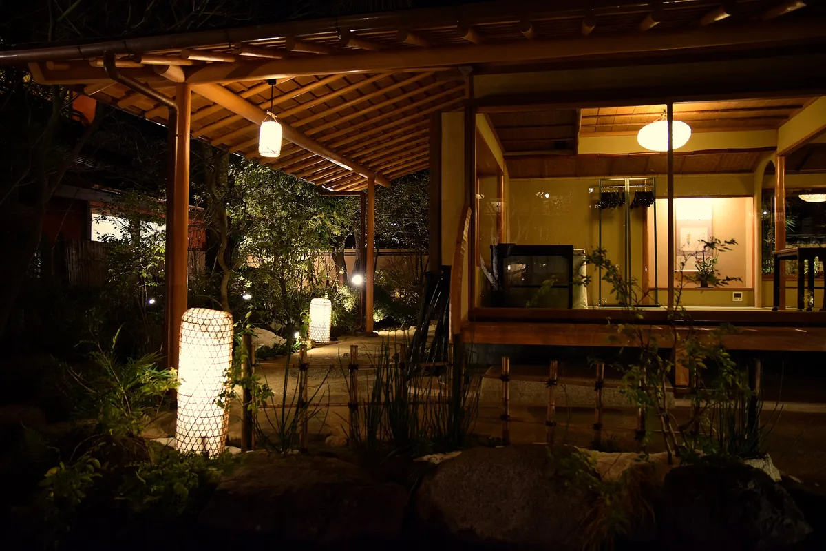 An evening garden with lit paper lanterns, a covered wooden veranda and a glowing tatami room beyond the glass