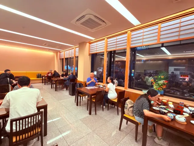 A brightly lit dining hall with wooden tables, diners and long windows along the wall
