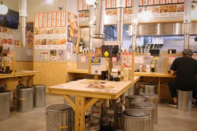 A plain eatery with wooden tables, metal buckets as stools and dish posters on the wall