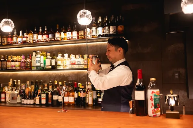 A bartender in a waistcoat shaking a cocktail shaker in front of lit bottle shelves