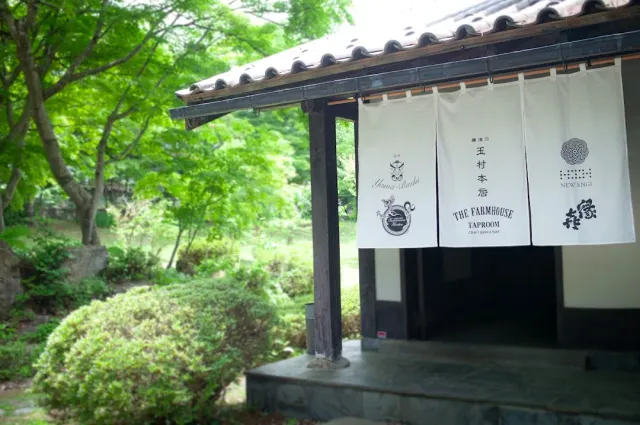 The entrance to a Japanese wooden house with white printed fabric curtains, surrounded by shrubs and green maples